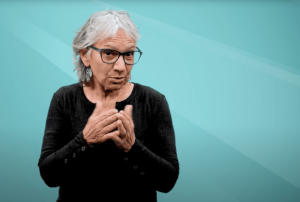 A white woman with grey hair and glasses, Dr. Gina Oliva, author, signs about enriching horizontal identities and deaf education that occurs outside the classroom.