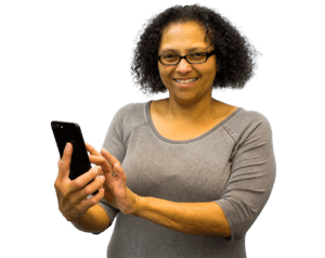 A woman with dark curly hair and glasses smiles into camera while wearing a grey shirt and holding a phone.