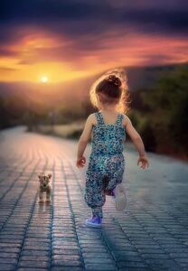 A girl toddler in a light blue romper chases a dog at sunset.