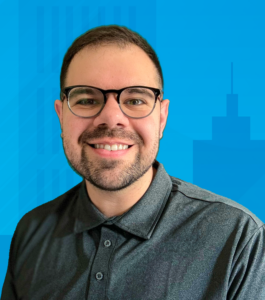 In the picture, a smiling white Hispanic cisgender man faces the camera. He has short dark hair, a beard, glasses, and a grey polo shirt. Behind him is a light blue background with abstract shapes resembling a city skyline in the lower right corner.
