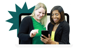 A white woman in a black blazer and green shirt looks at an IPhone with a black woman who is wearing a yellow top and a blazer. Both are smiling.