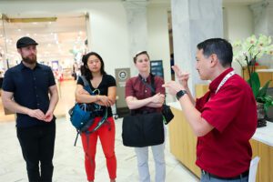 A dark-haired man wearing a red shirt and lanyard uses American Sign Language with three education administrators during a tour at the Administrator Summer Immersion Experience.