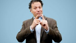 A man with curly dark hair wearing a blue oxford and brown blazer signs into camera about the Clerc Center's Administrator Summer Immersion Experience.