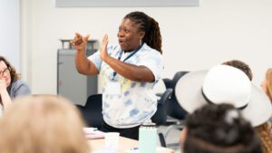A black woman in a tie-dye t-shirt signs to a group of administrators during Administrator Summer Immersion Experience week.