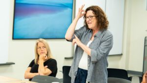 A Caucasian woman with auburn hair and glasses, wearing a grey dress shirt over a white top, signs to a group of deaf education administrators during Administrator Summer Immersion Experience week as part of the Clerc Center's School and State Partnership program.