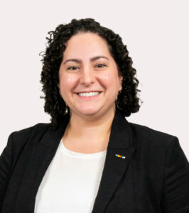 Sarah Honigfeld, a brunette woman with shoulder-length curly hair poses for her Clerc Center directory photo wearing a dark blazer and a white top. Honigfeld serves as the Director for Early Intervention.