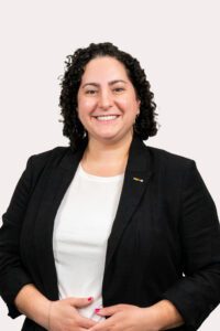 Sarah Honigfeld, a brunette woman with shoulder-length curly hair poses for her Clerc Center directory photo wearing a dark blazer and a white top. Honigfeld serves as the Director for Early Intervention.