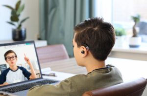 A deaf elementary-aged boy with dark hair and a hearing aid signs to a friend during a Zoom meeting. The friend, wearing glasses and a navy and white shirt, signs back to him.