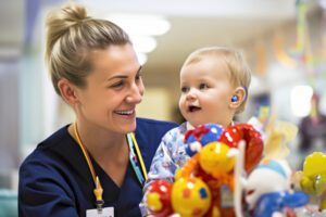 A Caucasian woman wearing navy scrubs with a badge and stethoscope around her neck smiles at a deaf infant.