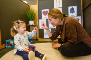 A blonde teacher wearing a brown sweater at Kendall Demonstration Elementary School (KDES) uses American Sign Language with a young blonde girl in pigtails. She girl is wearing a multi-colored blouse and is holding a purple picture book. The photo represents the early acquisition of language.