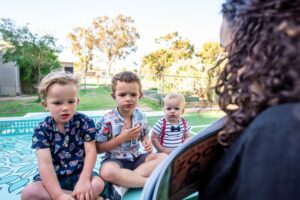 A woman with dark, curly hair is reading a book to three deaf or hard of hearing boys. The two boys on the left are in Pre-kindergarten and the third boy is a toddler. The middle boy in a short-sleeve button up shirt is using American Sign Language with the woman reading.