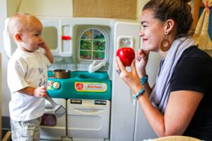 A young woman with brown hair wearing a black t-shirt and holding an apple signs and smiles at a deaf toddler in front of a play kitchen as part of Early Intervention training.