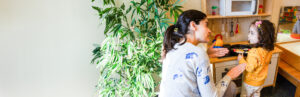 A woman with a dark ponytail is engaged with a deaf or hard of hearing toddler wearing an orange sweater in front of a play kitchen. The woman is using sign language. The photo represents early language acquisition.