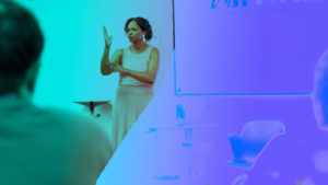 Barbara Robinson, Director of Clerc Center State and School Partnerships, signs to an audience of education administrators during Administrator Summer Immersion Experience. She is standing in front of a classroom whiteboard wearing a cream-colored top and long business skirt.