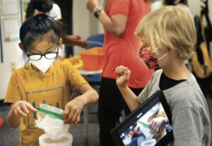 Two young deaf children wearing pandemic-era masks sign to each other while conducting a science experiment at elementary school.