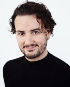 A smiling Persian person with medium-length, brown, curly hair and light skin, with a dimple on the left side. They have brown eyes and light facial hair, including a short beard and mustache. They are wearing a black high-neck sweater.
